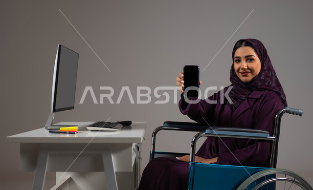 إتاحة المجال امام ذوي الهمم للدخول الى مجال العمل, تسهيل حياة المعاقين باستخدام التقنية والتكنولوجيا, بورتريه مقرب لشابة عربية خليجية سعودية مبتسمة ترتدي العباءة والحجاب تجلس على كرسي متحرك تقوم بعرض شاشة سوداء فارغه على الهاتف المحمول داخل المكتب, خلفية رمادية