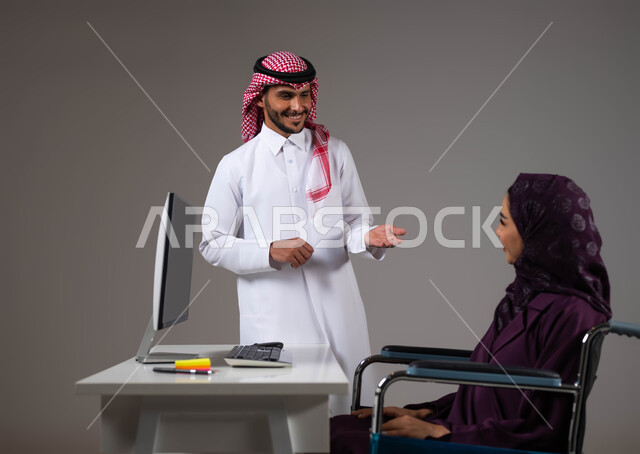 تبادل الافكار والخبرات لتحقيق اهداف الشركة، تمكين اصحاب الهمم وذوي الاحتياجات الخاصة من الدخول الى مجال العمل، بورتريه مقرب لزميلين عربيين خليجيين سعوديين يقومان بإنجاز المهام عبر الحاسوب المحمول داخل المكتب، دمج العمل مع التقنية والتكنولوجيا، خلفية رمادية