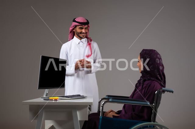 Listening to notes with love and passion, enabling people of determination and people with special needs to enter the field of work, a close-up portrait of two smiling Saudi Gulf Arab colleagues completing tasks via a laptop inside the office, integrating work with technology and technology, gray background
