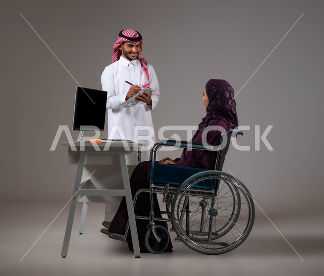 Writing and recording notes to develop the company’s plans, integrating work with technology and technology, providing the opportunity for people of determination to enter the field of work, a portrait of two smiling Gulf Arab Saudi colleagues completing tasks via a laptop inside the office, one team spirit, gray background