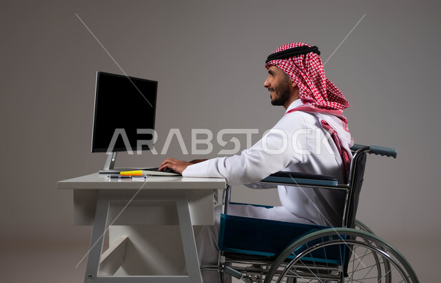 Providing opportunities for people of determination to enter the field of work, providing opportunities and jobs for the sick and disabled, a portrait of a smiling Gulf Arab Saudi man wearing a shemagh and traditional thobe, sitting in a wheelchair working on a computer at the company’s headquarters, social empowerment in Saudi society, gray background