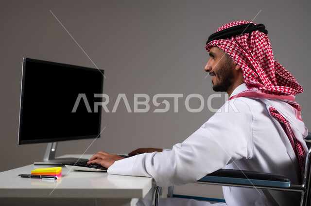 إتاحة المجال امام ذوي الهمم للدخول الى مجال العمل، توفير فرص ووظائف للمرضى والمعاقين، بورتريه مقرب لشاب عربي خليجي سعودي يرتدي الشماغ والثوب التقليدي يجلس على الكرسي المتحرك يعمل على جهاز الحاسوب في مقر الشركة, التمكين الاجتماعي في المجتمع السعودي، خلفية رمادية