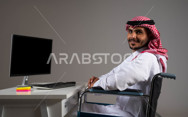 التمكين الاجتماعي في المجتمع السعودي، توفير بيئة عملية مريحة للمرضى والمعاقين، بورتريه لشاب عربي خليجي سعودي يرتدي الشماغ والثوب التقليدي يجلس على الكرسي المتحرك يعمل على جهاز الحاسوب في مقر الشركة, إتاحة المجال امام ذوي الهمم للدخول الى مجال العمل، خلفية رمادية