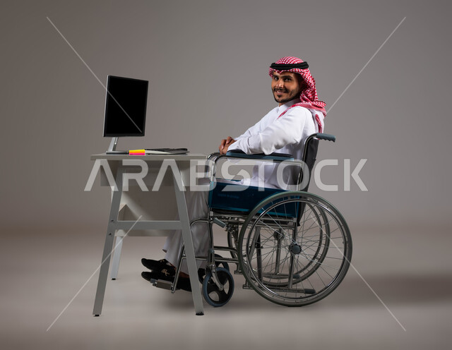 التمكين الاجتماعي في المجتمع السعودي، توفير بيئة عملية مريحة للمرضى والمعاقين، بورتريه لشاب عربي خليجي سعودي يرتدي الشماغ والثوب التقليدي يجلس على الكرسي المتحرك يعمل على جهاز الحاسوب في مقر الشركة, إتاحة المجال امام ذوي الهمم للدخول الى مجال العمل، خلفية رمادية