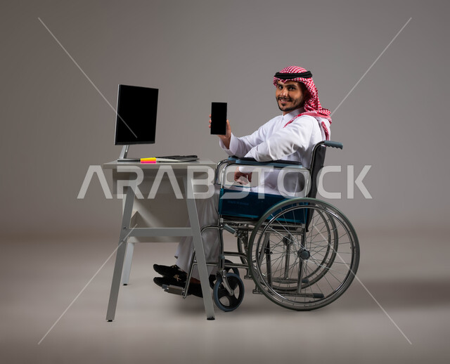 تسهيل حياة المعاقين باستخدام التقنيه والتكنولوجيا, شاشة سوداء فارغه على الهاتف المحمول, بورتريه لشاب عربي خليجي سعودي يرتدي الزي التقليدي يجلس على كرسي متحرك يقوم بعرض برامج ذوي الاحتياجات الخاصه, إتاحة المجال امام ذوي الهمم للدخول الى مجال العمل، خلفية رمادية
