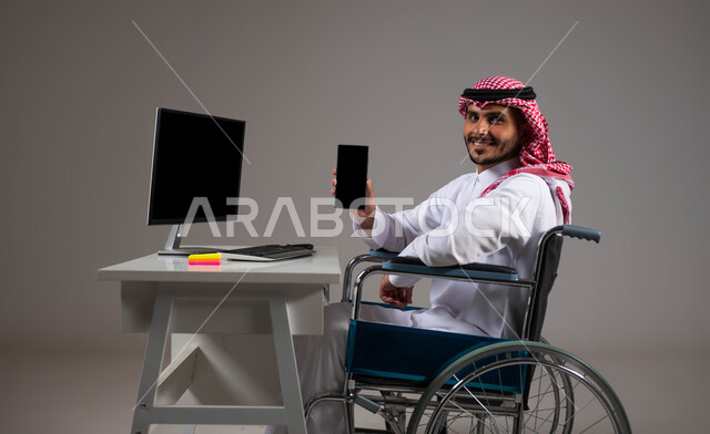 شاشة سوداء فارغه على الهاتف المحمول, بورتريه لشاب عربي خليجي سعودي يرتدي الزي التقليدي يجلس على كرسي متحرك يقوم بعرض برامج ذوي الاحتياجات الخاصه, إتاحة المجال امام ذوي الهمم للدخول الى مجال العمل، تسهيل حياة المعاقين باستخدام التقنيه والتكنولوجيا, خلفية رمادية