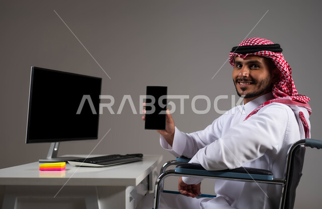 شاشة سوداء فارغه على الهاتف المحمول, بورتريه لشاب عربي خليجي سعودي يرتدي الزي التقليدي يجلس على كرسي متحرك يقوم بعرض برامج ذوي الاحتياجات الخاصه, إتاحة المجال امام ذوي الهمم للدخول الى مجال العمل، تسهيل حياة المعاقين باستخدام التقنيه والتكنولوجيا, خلفية رمادية