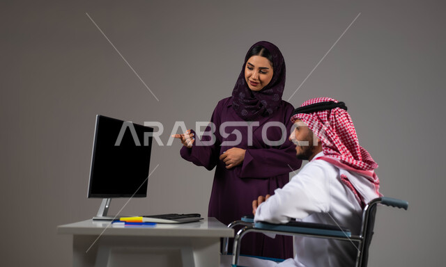 Pointing to the laptop and warning to implement plans, exchanging ideas and experiences to achieve goals, a portrait of two smiling Gulf Arab Saudi colleagues completing tasks via the laptop inside the office, integrating people of determination into work life, providing a suitable work environment for the disabled, gray background