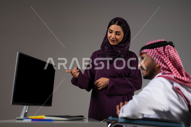 تبادل الأفكار والخبرات لتحقيق الأهداف، بورتريه لزميلين عربيين خليجيين سعوديين مبتسمين يقومان بإنجاز المهام عبر الحاسوب المحمول داخل المكتب، الاشارة الى اللابتوب والتنبيه لتطبيق الخطط، دمج ذوي الهمم بالحياة العملية، توفير بيئة عمل مناسبة للمعوقين، خلفيه رمادية