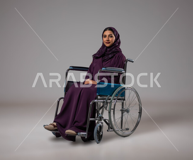 شعور المعاقين بالإنتماء والتقدير في السعوديه، العنايه بأصحاب الهمم وتأمين احتياجاتهم، بورتريه لإمرأة مريضة عربية سعودية خليجية مبتسمة ترتدي الحجاب والعباءة من ذوي الاحتياجات الخاصه تجلس على كرسي متحرك، توفير اجهزة ومعدات تساعد بالحركة والتنقل، خلفيه رمادية