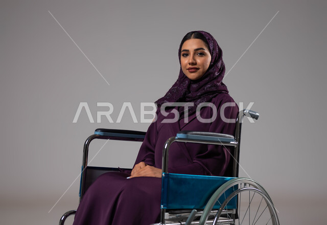 العنايه بأصحاب الهمم وتأمين احتياجاتهم، بورتريه لإمرأة مريضة عربية سعودية خليجية مبتسمة ترتدي الحجاب والعباءة من ذوي الاحتياجات الخاصه تجلس على كرسي متحرك، شعور المعاقين بالإنتماء والتقدير في السعوديه، توفير اجهزة ومعدات تساعد بالحركة والتنقل، خلفيه رمادية