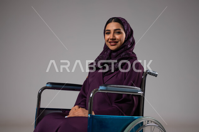 توفير اجهزة ومعدات تساعد بالحركة والتنقل، العنايه بأصحاب الهمم وتأمين احتياجاتهم، بورتريه مقرب لإمرأة مريضة عربية سعودية خليجية مبتسمة ترتدي الحجاب والعباءة من ذوي الاحتياجات الخاصه تجلس على كرسي متحرك، شعور المعاقين بالإنتماء والتقدير في السعوديه، خلفيه رمادية