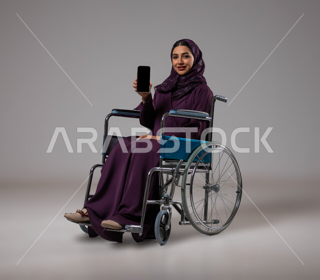 Facilitating the lives of the disabled using technology and technology, a blank black screen on the mobile phone, a portrait of a Saudi Gulf Arab young woman wearing the abaya and hijab, sitting in a wheelchair, displaying programs and applications for people with special needs, gray background