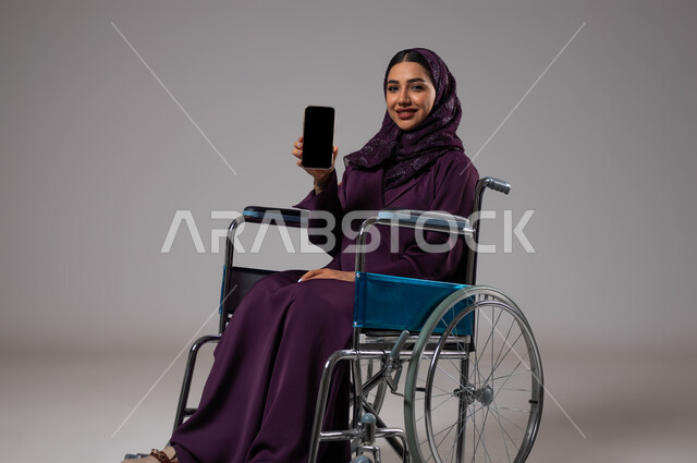 Facilitating the lives of the disabled using technology and technology, a blank black screen on the mobile phone, a portrait of a Saudi Gulf Arab young woman wearing the abaya and hijab, sitting in a wheelchair, displaying programs and applications for people with special needs, gray background