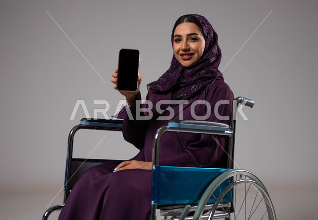 شاشة سوداء فارغه على الهاتف المحمول, بورتريه لشابة عربية خليجية سعودية ترتدي العباءة والحجاب تجلس على كرسي متحرك تقوم بعرض برامج وتطبيقات ذوي الاحتياجات الخاصه, تسهيل حياة المعاقين باستخدام التقنيه والتكنولوجيا, خلفية رمادية