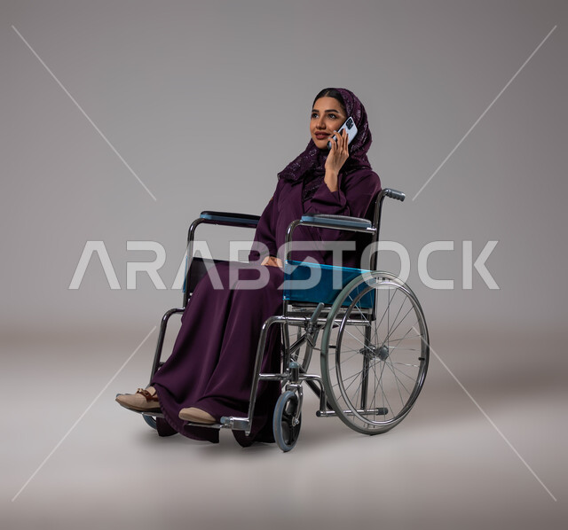 الدردشة والتحدث مع الاقارب عبر الهاتف المحمول, بورتريه لإمرأة عربية خليجية سعودية مبتسمة ترتدي الحجاب والعباءة تجلس على كرسي متحرك تقوم بإجراء مكالمة هاتفية, تأمين اجهزة ومعدات تساعد بالحركة والتنقل، قضاء وقت ممتع بالتواصل مع الاصدقاء, خلفية رمادية