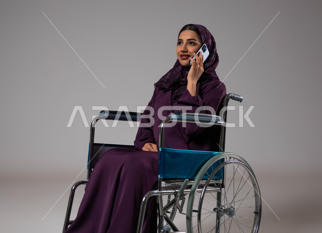 التحدث والدردشة مع الاقارب عبر الهاتف المحمول, بورتريه لإمرأة عربية خليجية سعودية مبتسمة ترتدي الحجاب والعباءة تجلس على كرسي متحرك تقوم بإجراء مكالمة هاتفية, تأمين اجهزة ومعدات تساعد بالحركة والتنقل، قضاء وقت ممتع بالتواصل مع الاصدقاء, خلفية رمادية