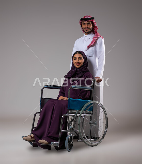 الوفاء والاخلاص بين الأزواج، تأمين معدات تساعد بالحركة والتنقل، بورتريه لزوج عربي خليجي سعودي يرتدي الزي السعودي يقوم بمساعدة زوجته من ذوي الاحتياجات الخاصه، مفهوم  العنايه ورعاية المرضى والمعاقين، الاهتمام بالجانب النفسي والجسدي للمقعدين، خلفية رمادية