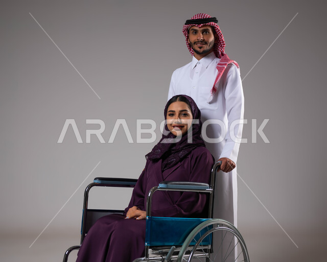 الاهتمام بالجانب النفسي والجسدي للمقعدين، الوفاء والاخلاص بين الأزواج، تأمين معدات تساعد بالحركة والتنقل، بورتريه لزوج عربي خليجي سعودي يرتدي الزي السعودي يقوم بمساعدة زوجته من ذوي الاحتياجات الخاصه، مفهوم  العنايه ورعاية المرضى والمعاقين، خلفية رمادية