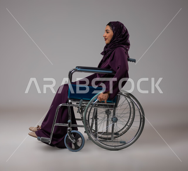 توفير اجهزة ومعدات تساعد بالحركة والتنقل، العنايه بأصحاب الهمم وتأمين احتياجاتهم الخاصة، بورتريه من الجانب لإمرأة مريضة عربية سعودية خليجية ترتدي الحجاب والعباءة تجلس على كرسي متحرك، شعور المعاقين بالإنتماء والتقدير والإحترام في السعوديه، خلفيه رمادية