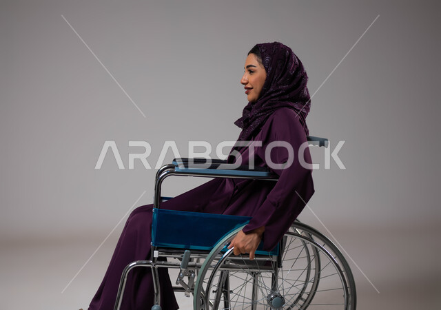 تأمين اجهزة ومعدات تساعد بالحركة والتنقل، العنايه بأصحاب الهمم وتأمين احتياجاتهم الخاصة، بورتريه من الجانب لإمرأة مريضة عربية سعودية خليجية ترتدي الحجاب والعباءة تجلس على كرسي متحرك، شعور المعاقين بالإنتماء والتقدير والإحترام في السعوديه، خلفيه رمادية