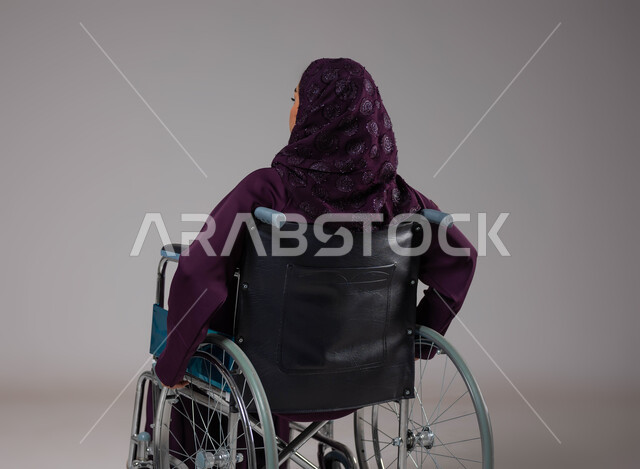 العنايه بأصحاب الهمم وتأمين احتياجاتهم الخاصة، بورتريه من الخلف لإمرأة مريضة عربية سعودية خليجية ترتدي الحجاب والعباءة تجلس على كرسي متحرك، شعور المعاقين بالإنتماء والتقدير والإحترام في السعوديه، توفير اجهزة ومعدات تساعد بالحركة والتنقل، خلفيه رمادية