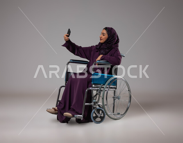 التقاط صور سيلفي تذكارية عبر الجوال، الاهتمام بأصحاب الهمم وتأمين احتياجاتهم الخاصة، بورتريه لإمرأة عربية سعودية خليجية ترتدي الحجاب والعباءة تجلس على كرسي متحرك تقوم باستخدام الهاتف المحمول، توفير اجهزة ومعدات تساعد بالحركة والتنقل، خلفيه رمادية