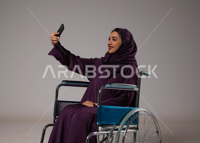 Taking souvenir selfies via mobile phone, caring for people of determination and providing for their special needs, a portrait of a Saudi Arabian Gulf woman wearing the hijab and abaya, sitting in a wheelchair using a mobile phone, providing devices and equipment that assist with movement and mobility, gray background.