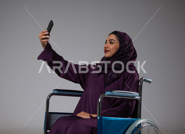 التقاط صور سيلفي تذكارية عبر الجوال، الاهتمام بأصحاب الهمم وتأمين احتياجاتهم الخاصة، بورتريه لإمرأة عربية سعودية خليجية ترتدي الحجاب والعباءة تجلس على كرسي متحرك تقوم باستخدام الهاتف المحمول، توفير اجهزة ومعدات تساعد بالحركة والتنقل، خلفيه رمادية