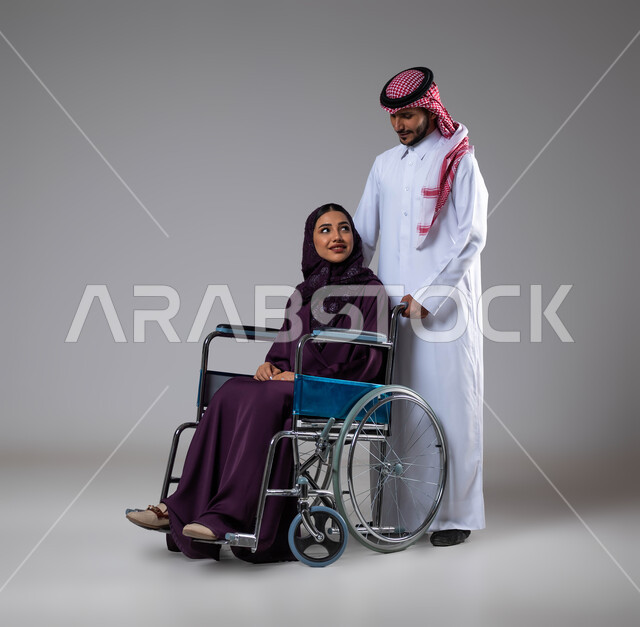المعامله الحسنه وتعابير البشاشه، الإخلاص  والعطف والحنان بين الأزواج، بورتريه لزوج عربي خليجي سعودي يرتدي الزي السعودي يقوم بمساعدة زوجته من ذوي الاحتياجات الخاصه، مفهوم  العنايه ورعاية المرضى والمعاقين، الاهتمام بالجانب النفسي والجسدي للمقعدين، خلفية رمادية