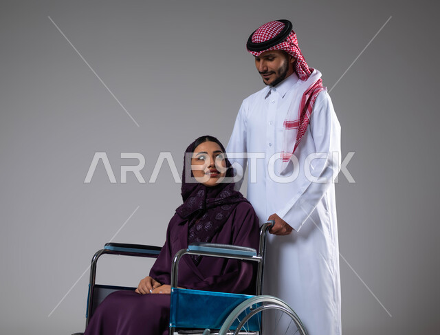 تعابير البشاشه والمعامله الحسنه، الإخلاص  والعطف والحنان بين الأزواج، بورتريه لزوج عربي خليجي سعودي يرتدي الزي السعودي يقوم بمساعدة زوجته من ذوي الاحتياجات الخاصه، مفهوم  العنايه ورعاية المرضى والمعاقين، الاهتمام بالجانب النفسي والجسدي للمقعدين، خلفية رمادية