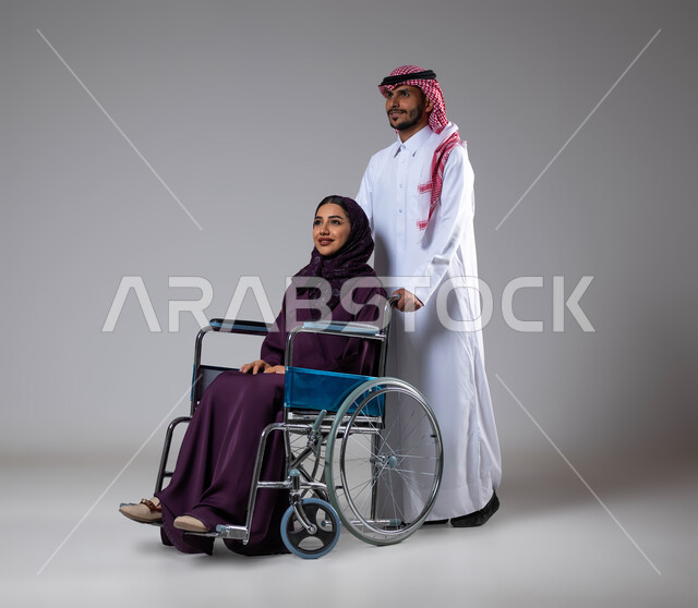 الإخلاص  والعطف والحنان بين الأزواج، تأمين معدات تساعد بالحركة والتنقل، بورتريه لزوج عربي خليجي سعودي يرتدي الزي السعودي يقوم بمساعدة زوجته من ذوي الاحتياجات الخاصه، مفهوم  العنايه ورعاية المرضى والمعاقين، الاهتمام بالجانب النفسي والجسدي للمقعدين، خلفية رمادية