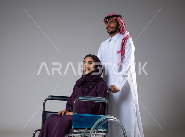 الوفاء والاخلاص بين الأزواج، تأمين معدات تساعد بالحركة والتنقل، بورتريه لزوج عربي خليجي سعودي يرتدي الزي السعودي يقوم بمساعدة زوجته من ذوي الاحتياجات الخاصه، مفهوم  العنايه ورعاية المرضى والمعاقين، الاهتمام بالجانب النفسي والجسدي للمقعدين، خلفية رمادية
