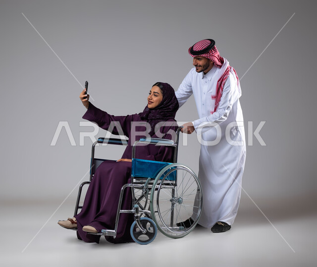 التقاط صور سيلفي تذكارية باستخدام الجوال، تأمين معدات تساعد بالحركة والتنقل، بورتريه لزوج عربي خليجي سعودي يرتدي الزي السعودي يقوم بمساعدة زوجته من ذوي الاحتياجات الخاصه، رعاية المرضى والمعاقين، الاهتمام بالجانب النفسي والجسدي للمقعدين، خلفية رمادية