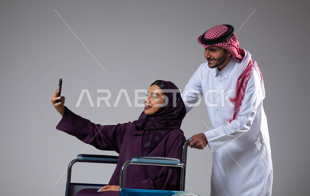 Providing equipment that helps with movement and movement, taking souvenir selfies using a mobile phone, a portrait of a Saudi Gulf Arab husband wearing the Saudi uniform helping his wife with special needs, caring for the sick and disabled, paying attention to the psychological and physical aspects of the disabled, gray background