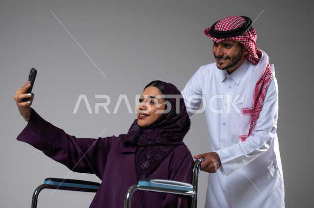 Providing equipment that helps with movement and movement, taking souvenir selfies using a mobile phone, a portrait of a Saudi Gulf Arab husband wearing the Saudi uniform helping his wife with special needs, caring for the sick and disabled, paying attention to the psychological and physical aspects of the disabled, gray background