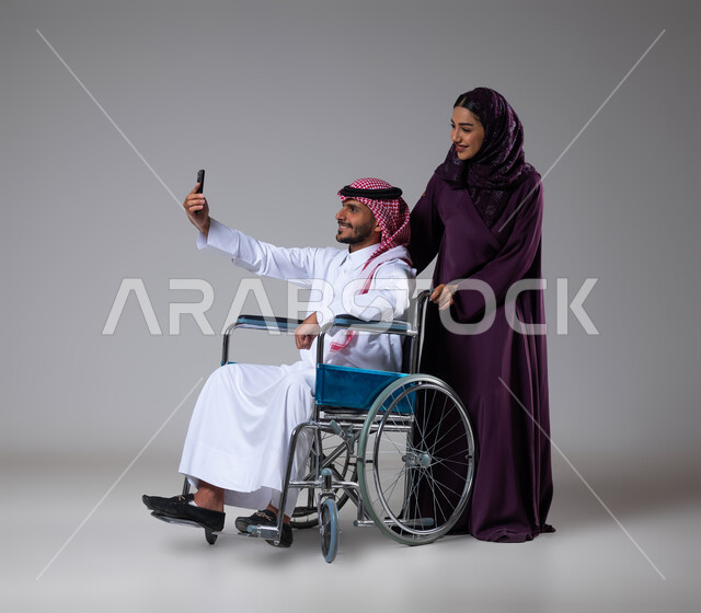 تأمين اجهزة ومعدات تساعد بالحركة والتنقل، العنايه والاهتمام بأصحاب الهمم، التقاط صور سيلفي تذكارية باستخدام الجوال، الإخلاص والحنان بين الأزواج، بورتريه لزوجة عربية خليجية سعودية محجبة ترتدي العباءة تقوم بمساعدة زوجها من ذوي الاحتياجات الخاصه، خلفية رمادية