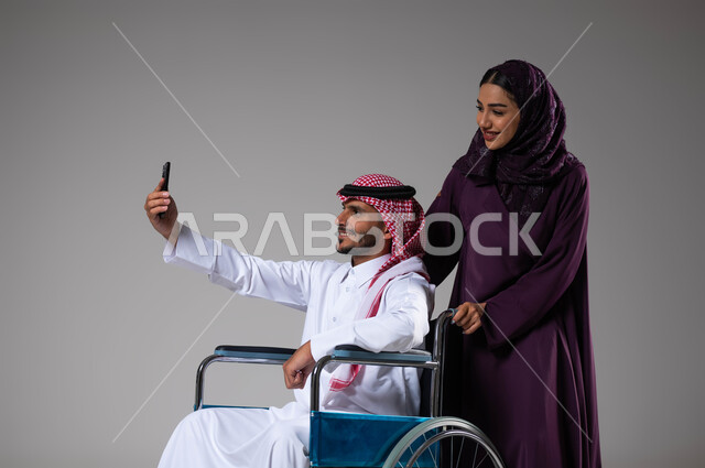 العنايه والاهتمام بأصحاب الهمم، التقاط صور سيلفي تذكارية باستخدام الجوال، الإخلاص والحنان بين الأزواج، بورتريه مقرب لزوجة عربية خليجية سعودية محجبة ترتدي العباءة تقوم بمساعدة زوجها من ذوي الاحتياجات الخاصه، تأمين اجهزة ومعدات تساعد بالحركة والتنقل، خلفية رمادية