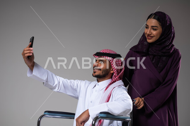 العنايه والاهتمام بأصحاب الهمم، التقاط صور سيلفي تذكارية باستخدام الجوال، الإخلاص والحنان بين الأزواج، بورتريه مقرب لزوجة عربية خليجية سعودية محجبة ترتدي العباءة تقوم بمساعدة زوجها من ذوي الاحتياجات الخاصه، تأمين اجهزة ومعدات تساعد بالحركة والتنقل، خلفية رمادية