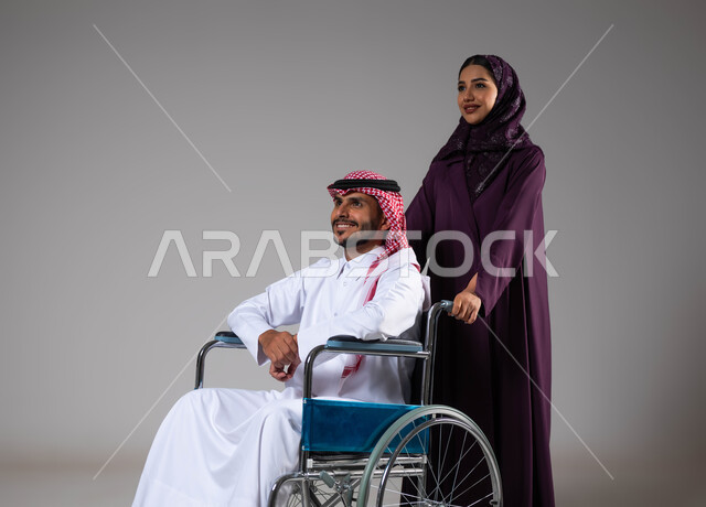 الحنان والإخلاص بين الأزواج, شعور المعاقين بالإنتماء والتقدير في السعوديه، بورتريه لزوجة عربية خليجية سعودية محجبة ترتدي العباءة تقوم بمساعدة زوجها من ذوي الاحتياجات الخاصه، تأمين اجهزة ومعدات تساعد بالحركة والتنقل، العنايه والاهتمام بأصحاب الهمم، خلفية رمادية