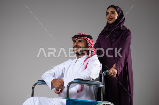 شعور المعاقين بالإنتماء والتقدير في السعوديه، بورتريه لزوجة عربية خليجية سعودية محجبة ترتدي العباءة تقوم بمساعدة زوجها من ذوي الاحتياجات الخاصه، تأمين اجهزة ومعدات تساعد بالحركة والتنقل، العنايه والاهتمام بأصحاب الهمم، الحنان والإخلاص بين الأزواج, خلفية رمادية