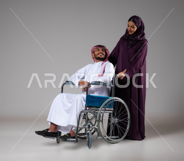 شعور المعاقين بالإنتماء والتقدير في السعوديه، بورتريه لزوجة عربية خليجية سعودية محجبة ترتدي العباءة تقوم بمساعدة زوجها من ذوي الاحتياجات الخاصه، تأمين اجهزة ومعدات تساعد بالحركة والتنقل، العنايه والاهتمام بأصحاب الهمم، الحنان والإخلاص بين الأزواج, خلفية رمادية