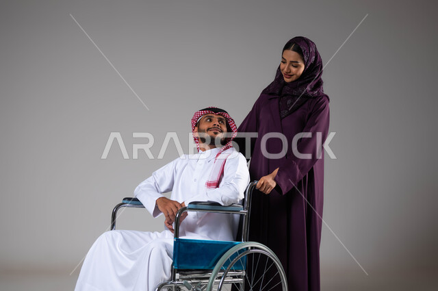 الإخلاص والحنان بين الأزواج, شعور المعاقين بالإنتماء والتقدير في السعوديه، بورتريه لزوجة عربية خليجية سعودية محجبة ترتدي العباءة تقوم بمساعدة زوجها من ذوي الاحتياجات الخاصه، تأمين اجهزة ومعدات تساعد بالحركة والتنقل، العنايه والاهتمام بأصحاب الهمم، خلفية رمادية