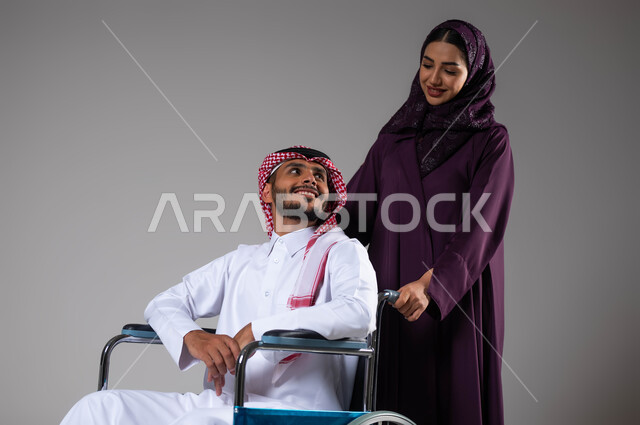 الإخلاص والحنان بين الأزواج, شعور المعاقين بالإنتماء والتقدير في السعوديه، بورتريه لزوجة عربية خليجية سعودية محجبة ترتدي العباءة تقوم بمساعدة زوجها من ذوي الاحتياجات الخاصه، تأمين اجهزة ومعدات تساعد بالحركة والتنقل، العنايه والاهتمام بأصحاب الهمم، خلفية رمادية
