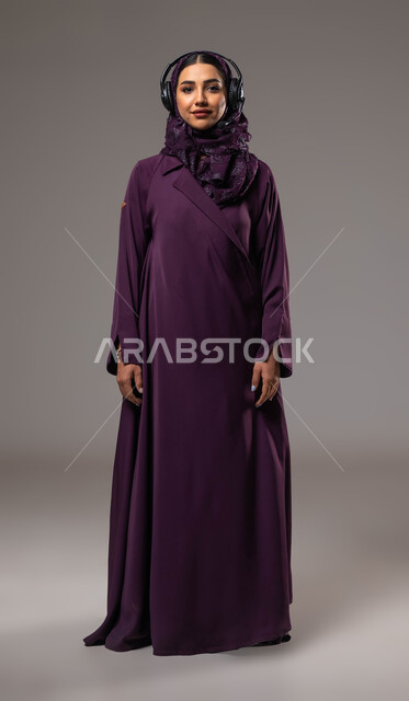 استخدام التقنيات المتطورة في العمل, الإجابة والرد على الاستفسارات والاستعلامات, التواصل مع الزبائن لحل المشكلات والشكاوي، بورتريه لامرأة عربية خليجية سعودية مبتسمة محجبة ترتدي العباءة تضع سماعات الرأس تعمل في قسم خدمة العملاء، صورة الجسم بالكامل، خلفية رمادية