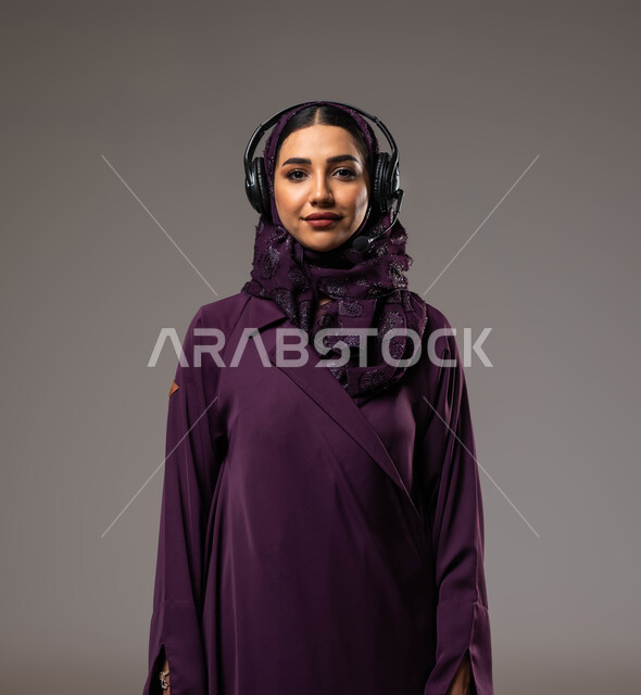 الإجابة والرد على الاستفسارات والاستعلامات, التواصل مع الزبائن لحل المشكلات والشكاوي، استخدام التقنيات المتطورة في العمل, بورتريه مقرب لامرأة عربية خليجية سعودية مبتسمة محجبة ترتدي العباءة تضع سماعات الرأس تعمل في قسم خدمة العملاء، خلفية رمادية