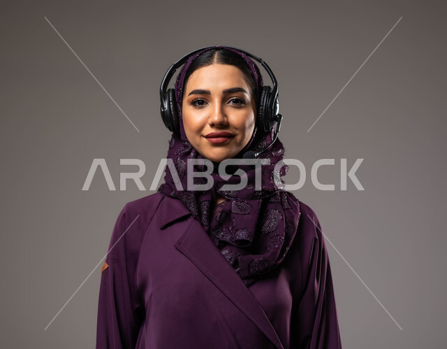 التواصل مع الزبائن لحل المشكلات والشكاوي، الإجابة والرد على الاستفسارات والاستعلامات, استخدام التقنيات المتطورة في العمل, بورتريه مقرب لامرأة عربية خليجية سعودية مبتسمة محجبة ترتدي العباءة تضع سماعات الرأس تعمل في قسم خدمة العملاء، خلفية رمادية