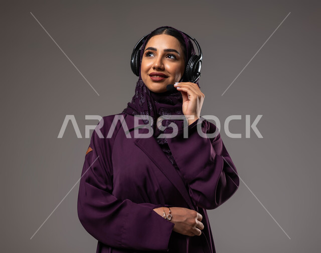 التواصل مع الزبائن لحل المشكلات والشكاوي، الإجابة والرد على الاستفسارات والاستعلامات, استخدام التقنيات المتطورة في العمل, بورتريه مقرب لامرأة عربية خليجية سعودية مبتسمة محجبة ترتدي العباءة تضع سماعات الرأس تعمل في قسم خدمة العملاء، خلفية رمادية