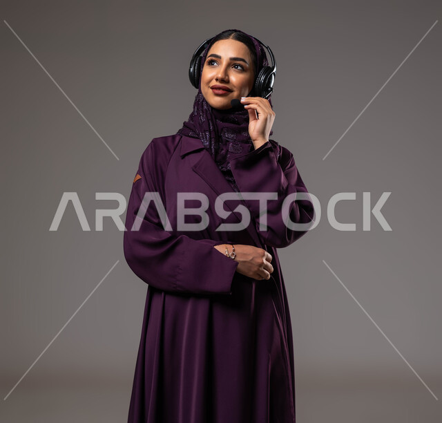 الإجابة والرد على الاستفسارات والاستعلامات, التواصل مع الزبائن لحل المشكلات والشكاوي، استخدام التقنيات المتطورة في العمل, بورتريه مقرب لامرأة عربية خليجية سعودية مبتسمة محجبة ترتدي العباءة تضع سماعات الرأس تعمل في قسم خدمة العملاء، خلفية رمادية