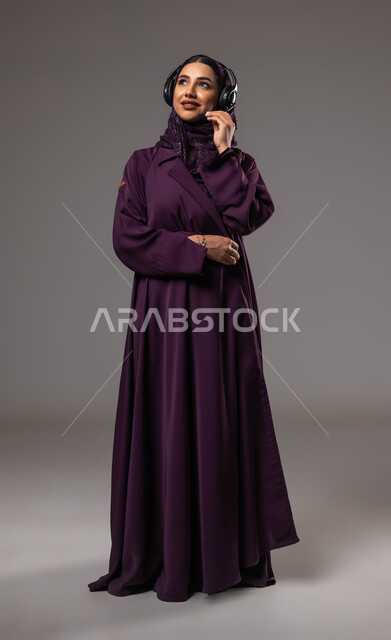 Using advanced technologies at work, answering and responding to inquiries and inquiries, communicating with customers to solve problems and complaints, portrait of a smiling Saudi Arabian Gulf woman, wearing a veil, wearing an abaya, wearing headphones, working in the customer service department, full body portrait, gray background